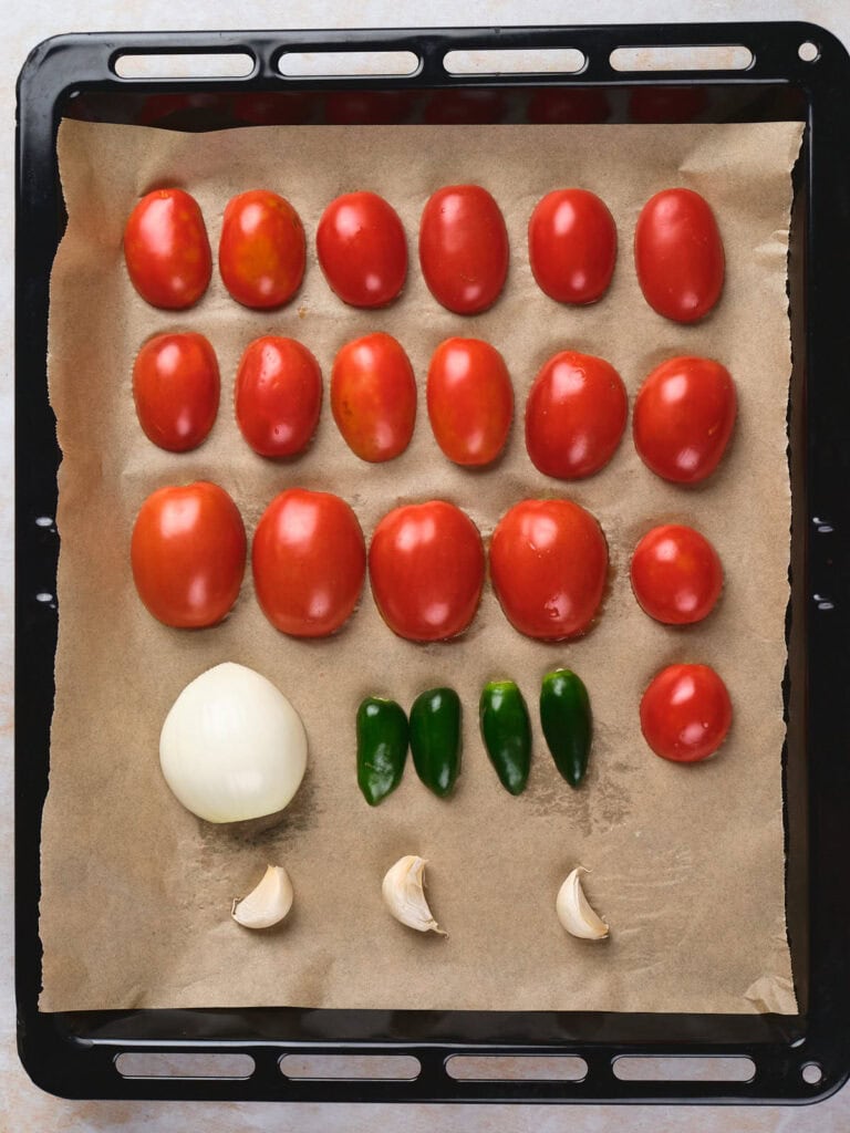 Assorted tomatoes, a whole onion, jalapeño peppers, and garlic cloves neatly arranged.