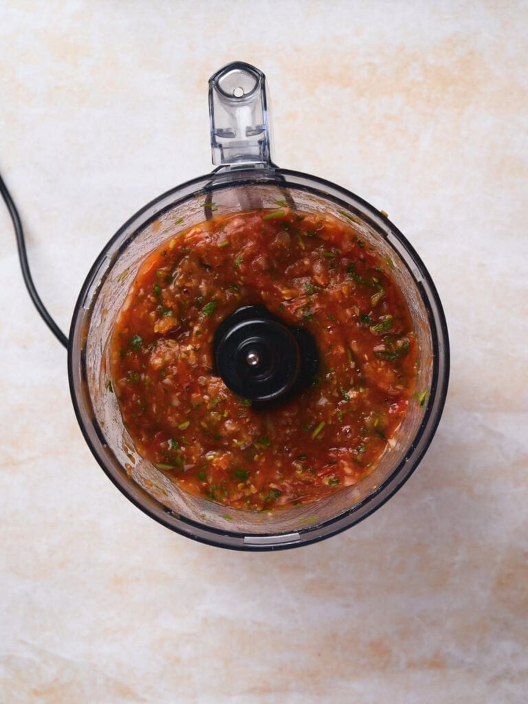 Top view of a food processor containing freshly blended tomato salsa with herbs.