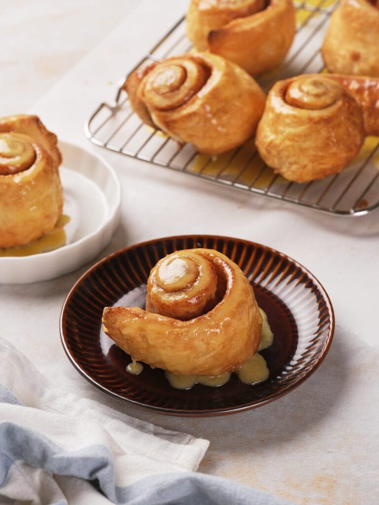 A glazed puff pastry cinnamon roll on a brown plate with additional rolls.