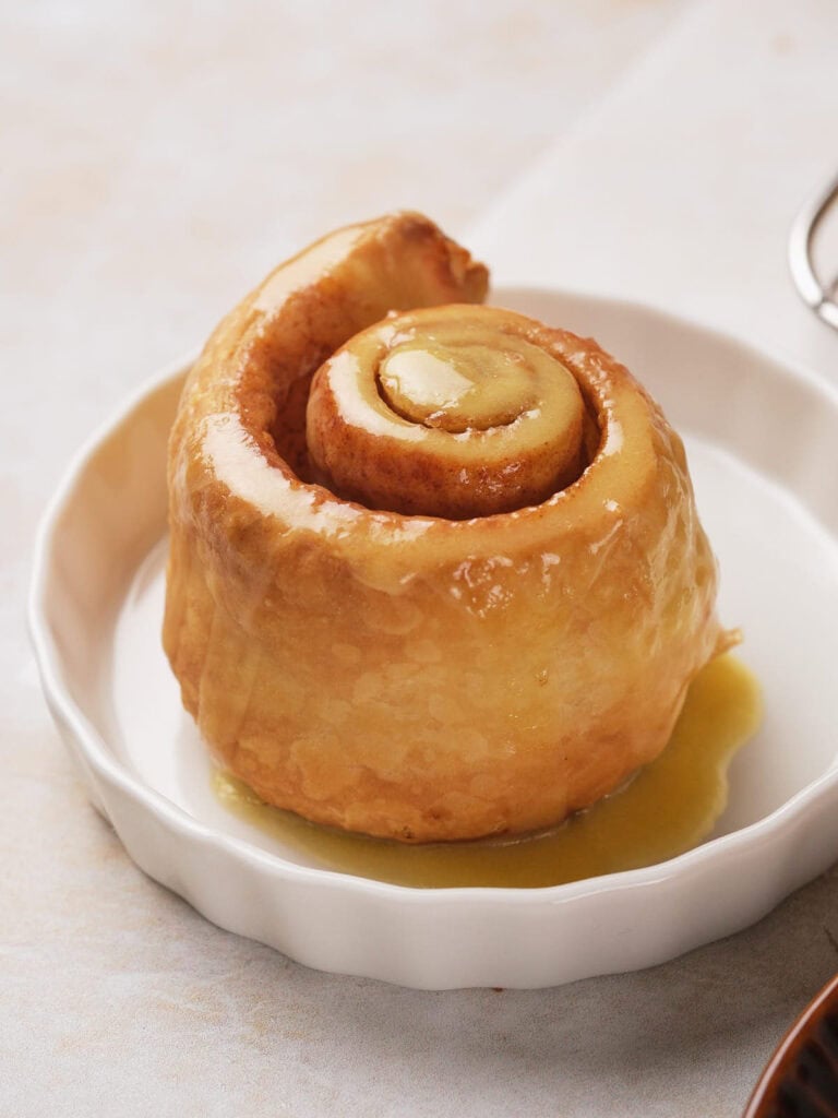 A glazed puff pastry cinnamon roll sits in a round white dish on a light surface.