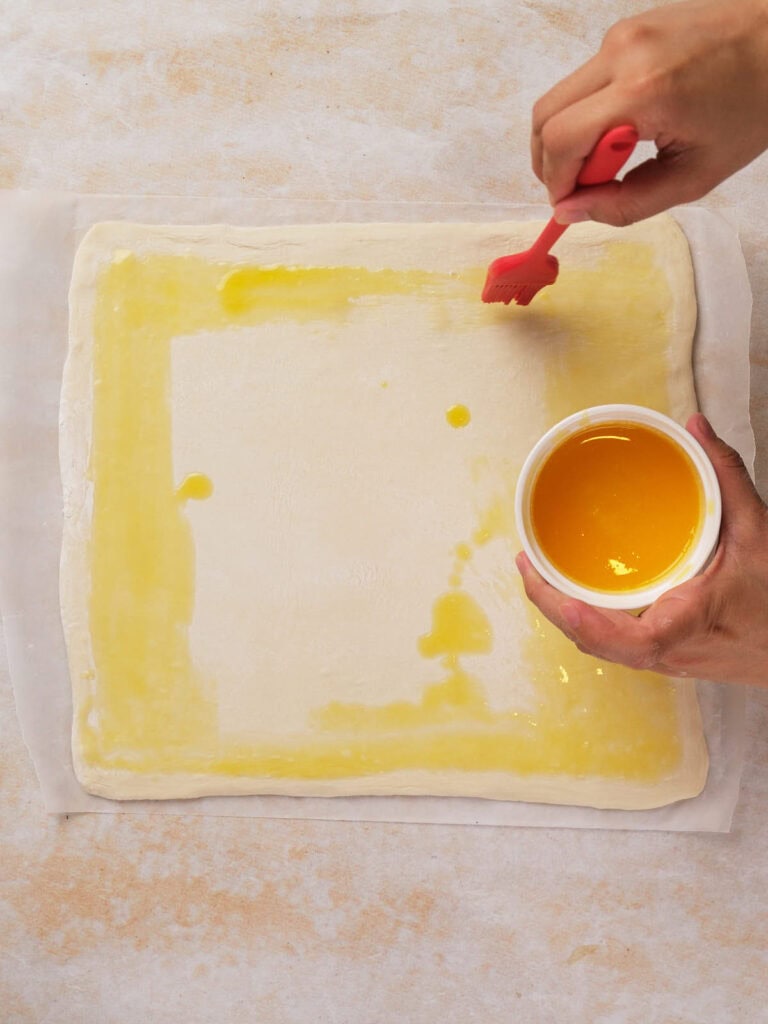 Hands brushing egg wash on a sheet of dough with a red brush.