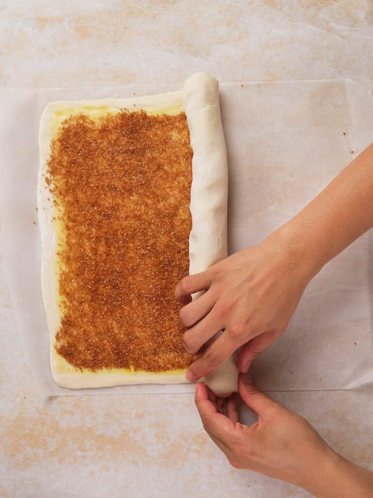 Hands rolling dough filled with a cinnamon-sugar mixture on parchment paper.