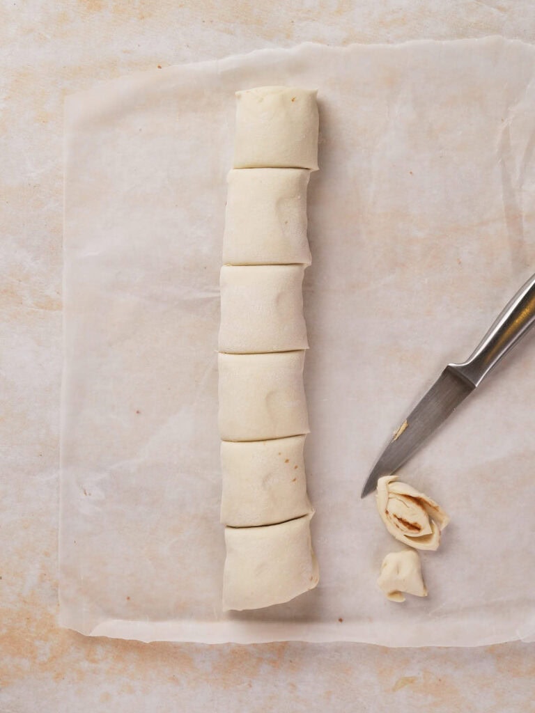 Seven evenly sliced pieces of rolled dough on parchment paper with a knife nearby.