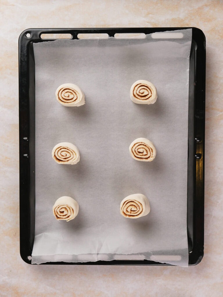 Six unbaked cinnamon rolls arranged evenly on a baking tray lined with parchment paper.