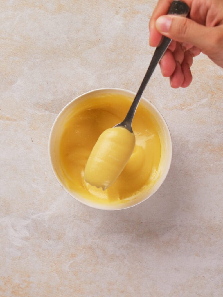 A hand holds a spoon with a smooth, yellow custard-like substance over a small white bowl on a light textured surface.