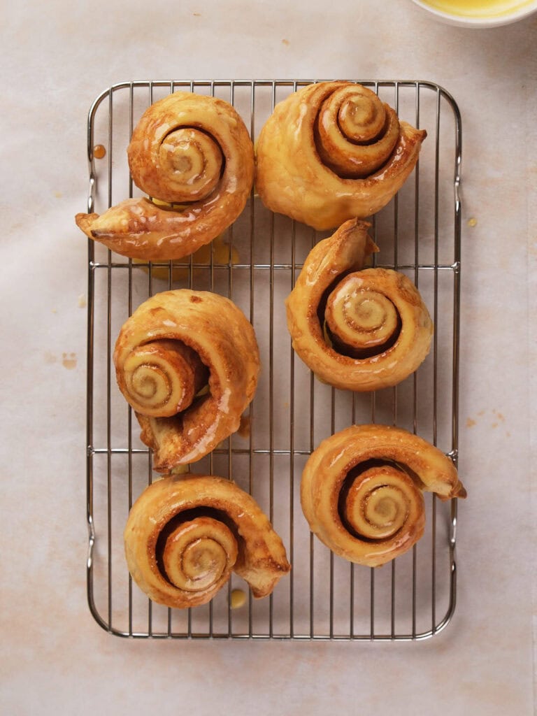 Six glazed cinnamon rolls on a wire rack on a light surface.