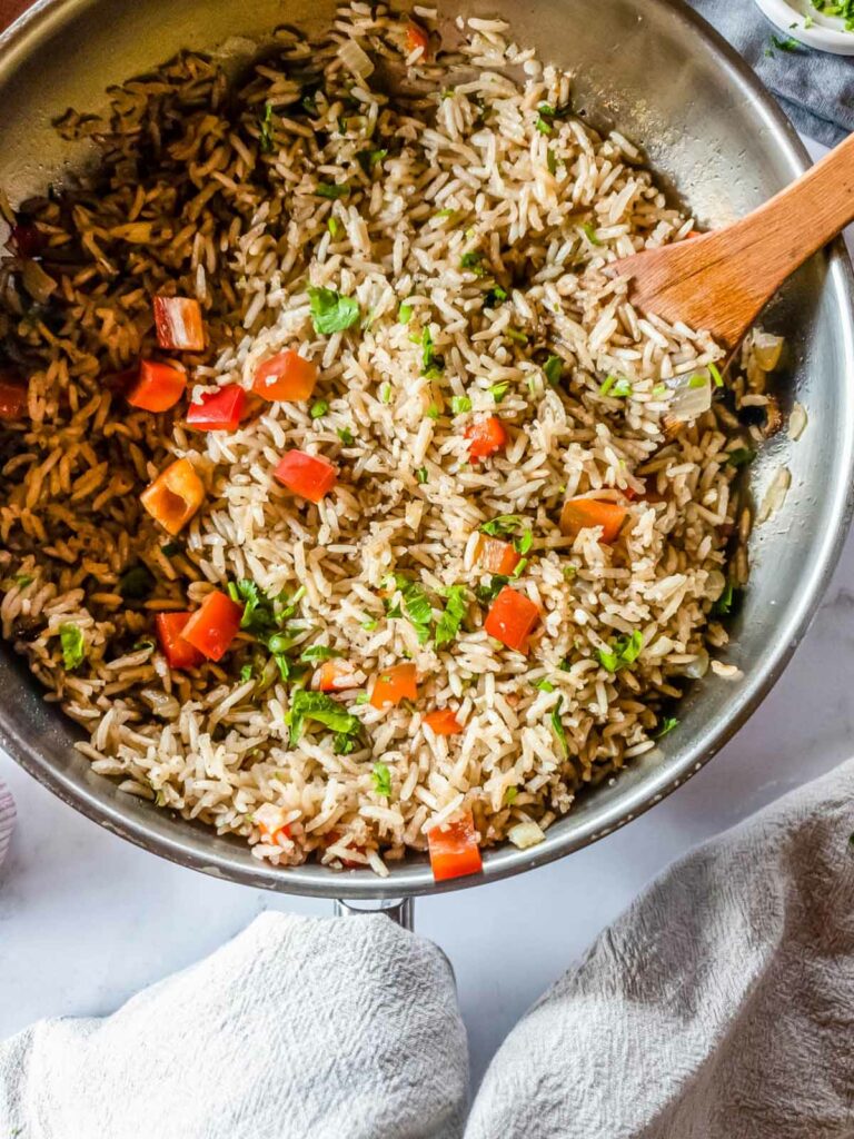 A pan of rice pilaf mixed with chopped red bell peppers and herbs, stirred with a wooden spoon.