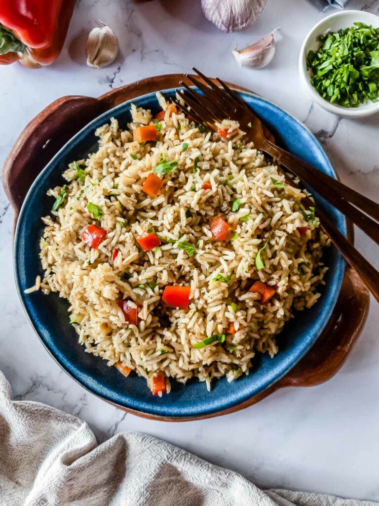 A plate of rice pilaf with red bell peppers and herbs, positioned on a marble surface next to a wooden fork and spoon.