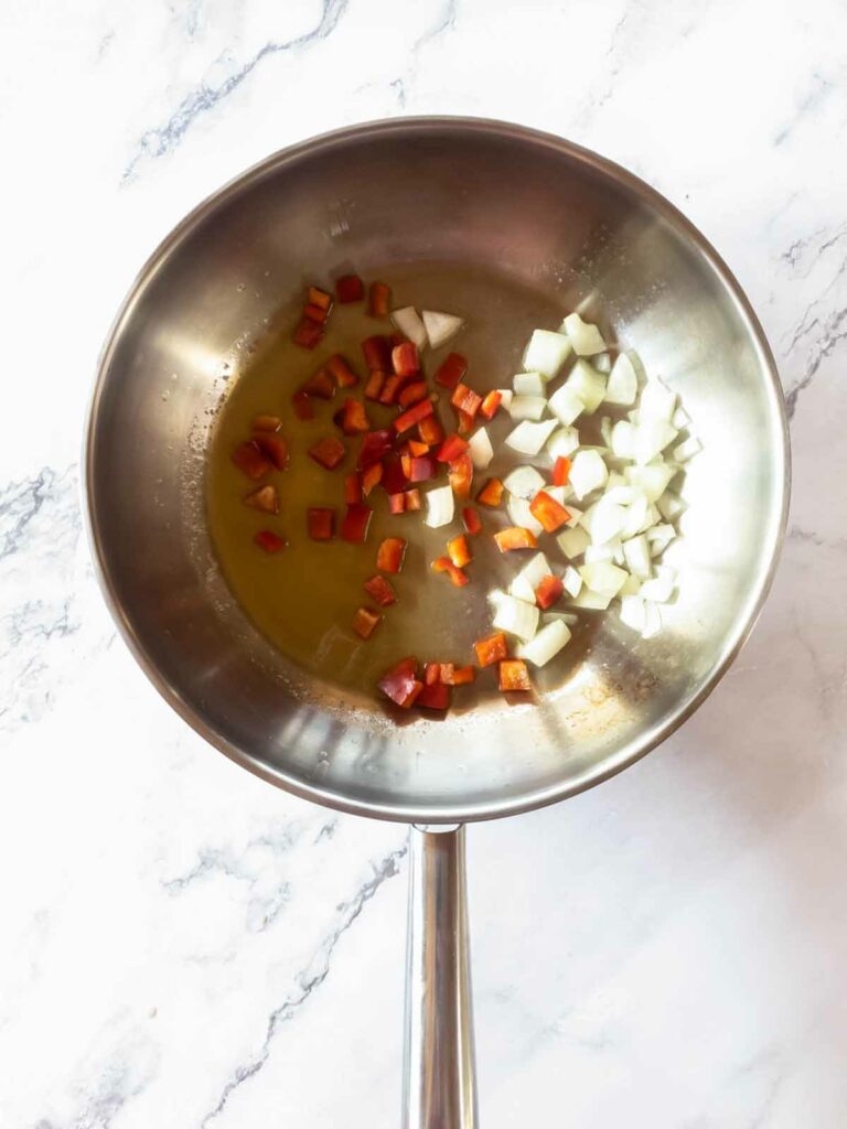 Chopped onions and red bell peppers in a stainless steel pan on a marble surface.