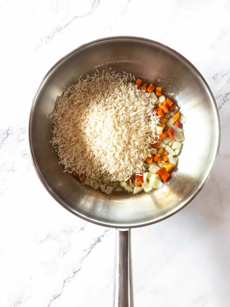 A stainless steel pan on a marble surface contains uncooked rice, chopped carrots, and diced onions.