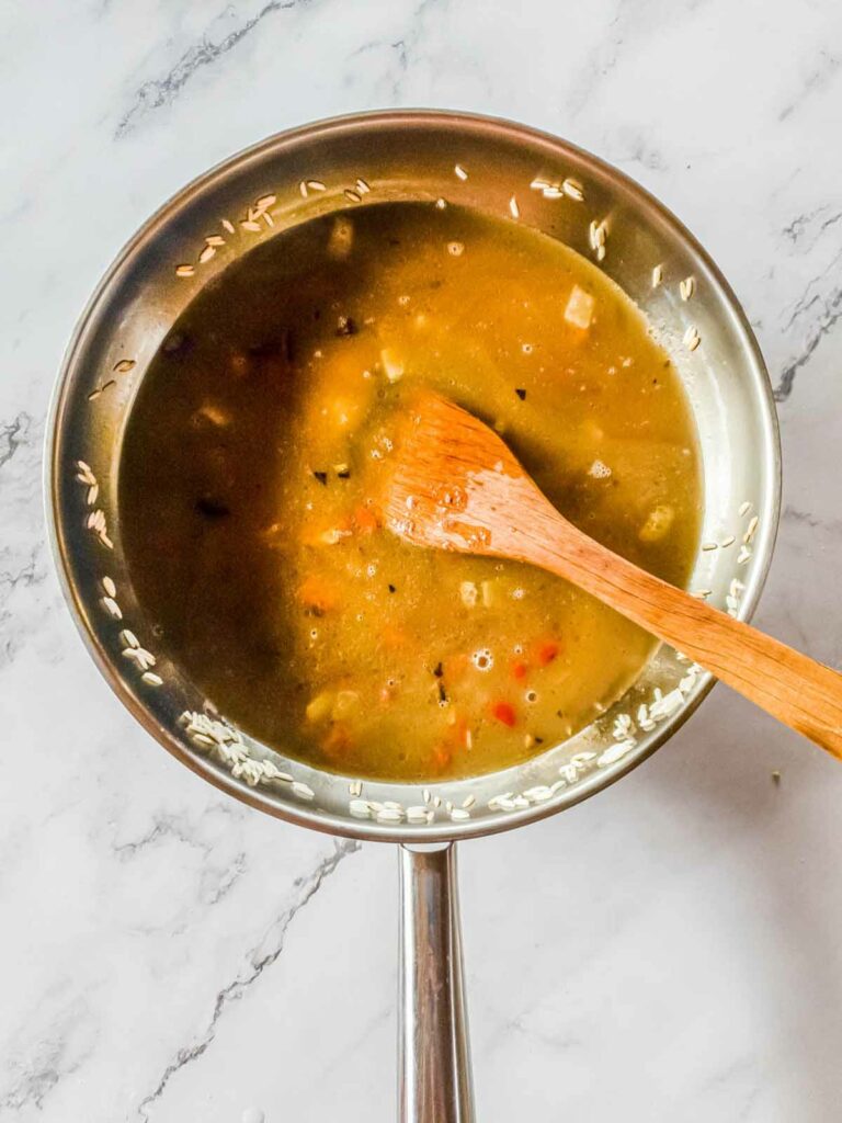 A saucepan with soup containing vegetables and a wooden spoon on a marble surface.