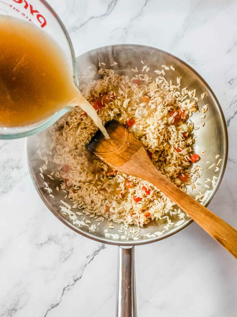 A measuring cup pouring liquid into a pan with rice and chopped vegetables being stirred with a wooden spoon.