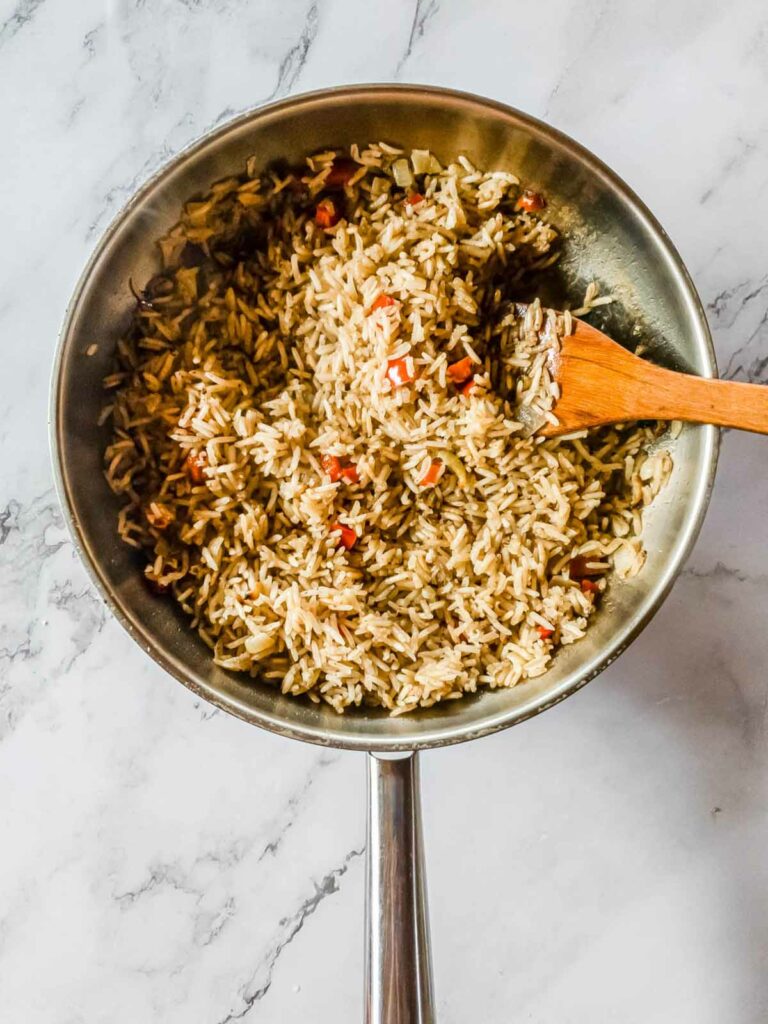 Pan with cooked rice, diced red bell peppers, and sliced mushrooms on a marble surface, stirred with a wooden spoon.