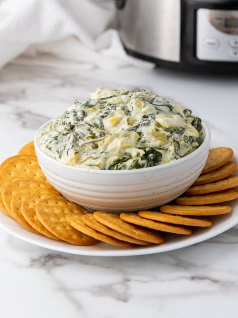 A bowl of creamy spinach and artichoke dip is surrounded by round crackers.