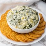 A bowl of spinach artichoke dip surrounded by a ring of round crackers on a white plate.