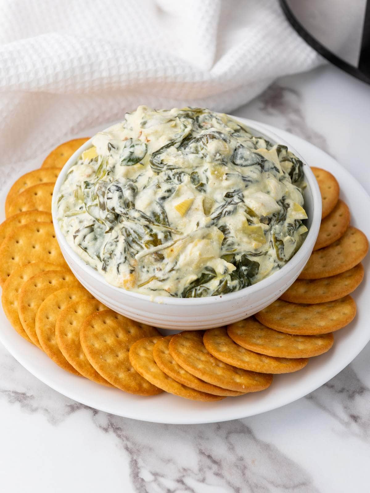 A bowl of spinach artichoke dip surrounded by a ring of round crackers on a white plate.