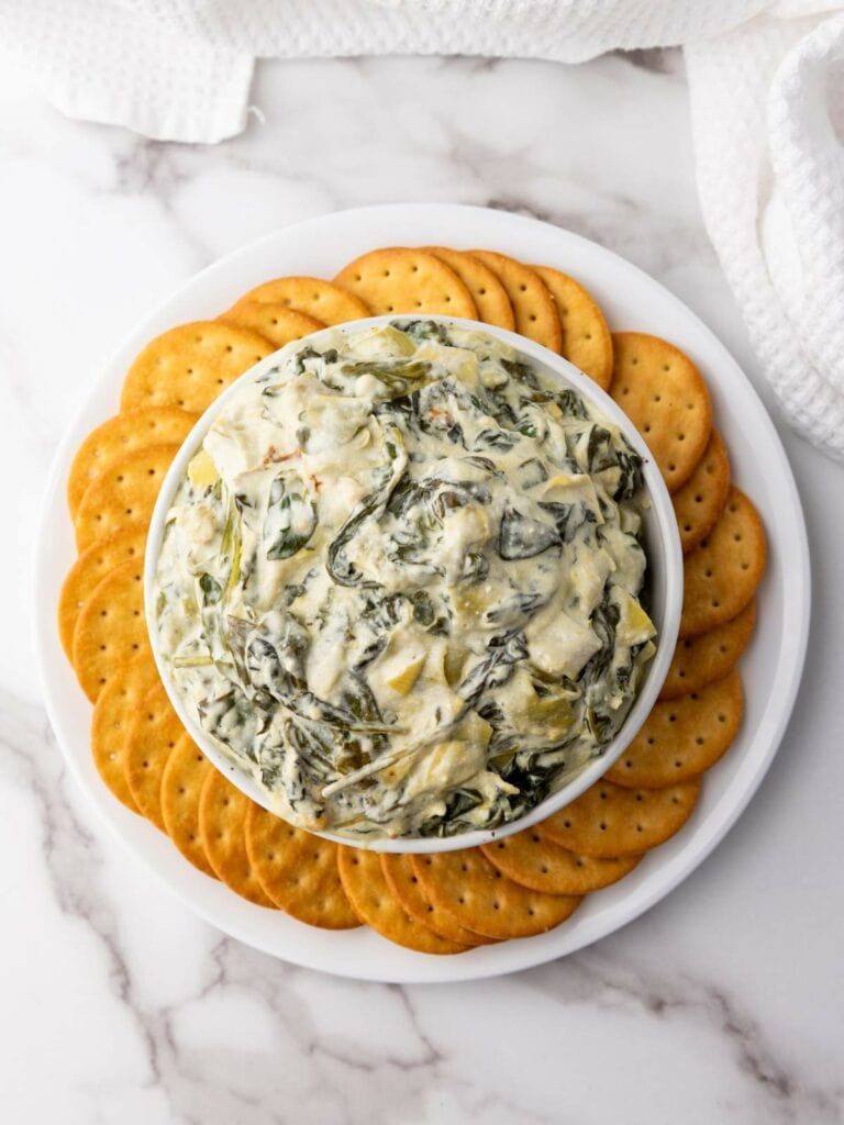 A bowl of creamy spinach artichoke dip surrounded by crackers on a white plate, set on a marble surface.