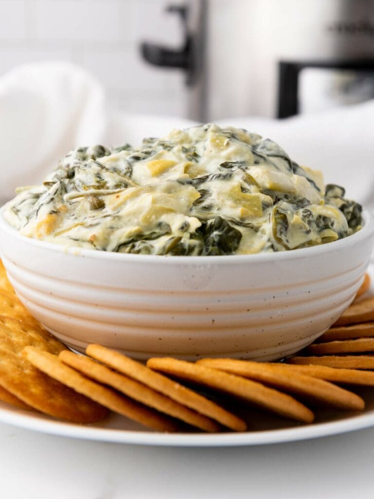 A bowl of creamy spinach artichoke dip surrounded by crackers on a white plate.