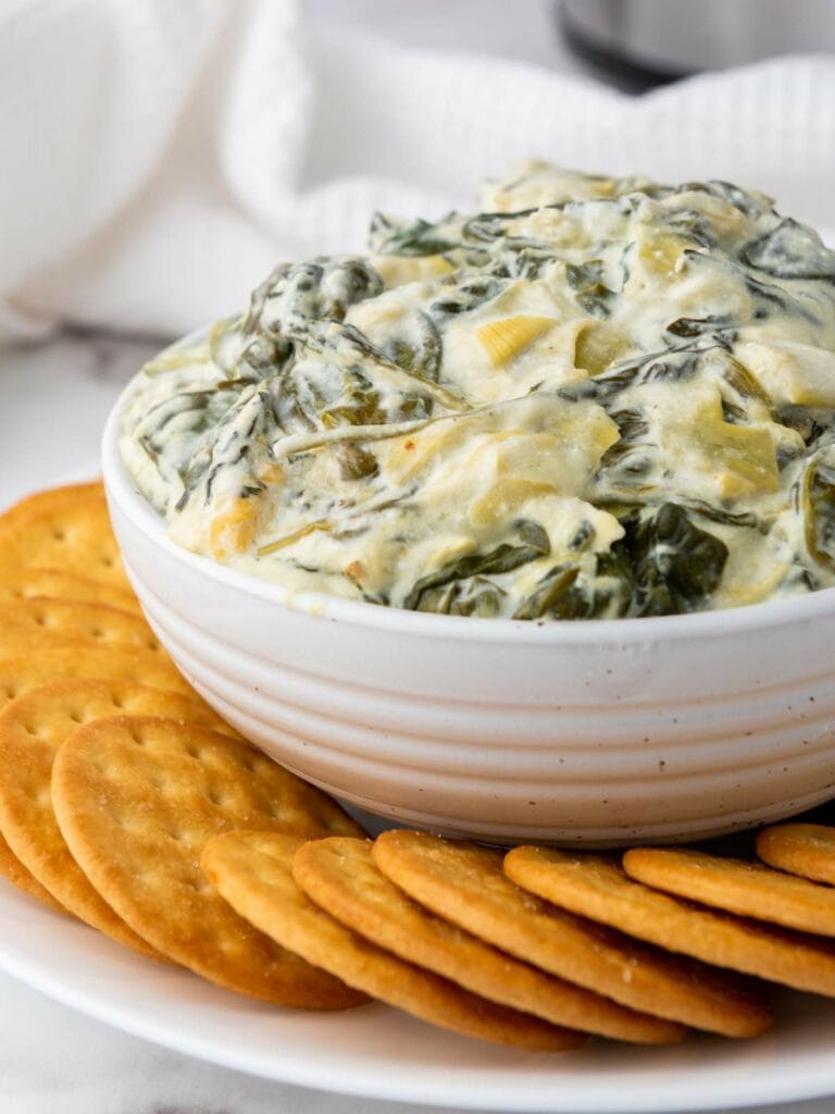 A bowl of creamy spinach and artichoke dip surrounded by a ring of crackers on a white plate.