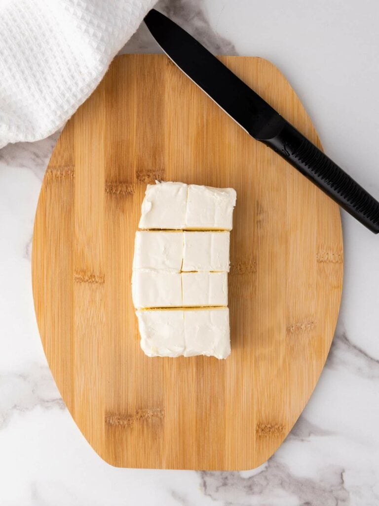 A knife and a white cloth are beside square pieces of frosted cake on a wooden cutting board.