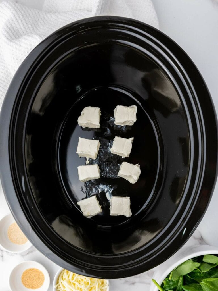 Black slow cooker with evenly spaced cream cheese cubes inside.