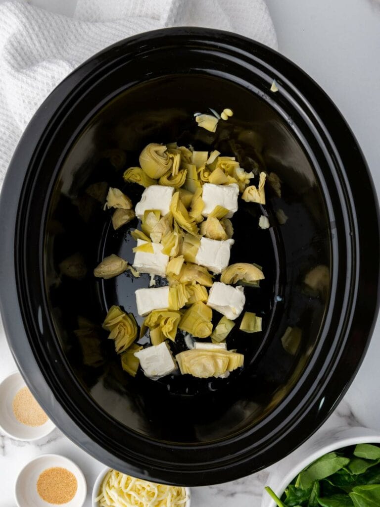 Artichoke hearts and cream cheese cubes in a black slow cooker.