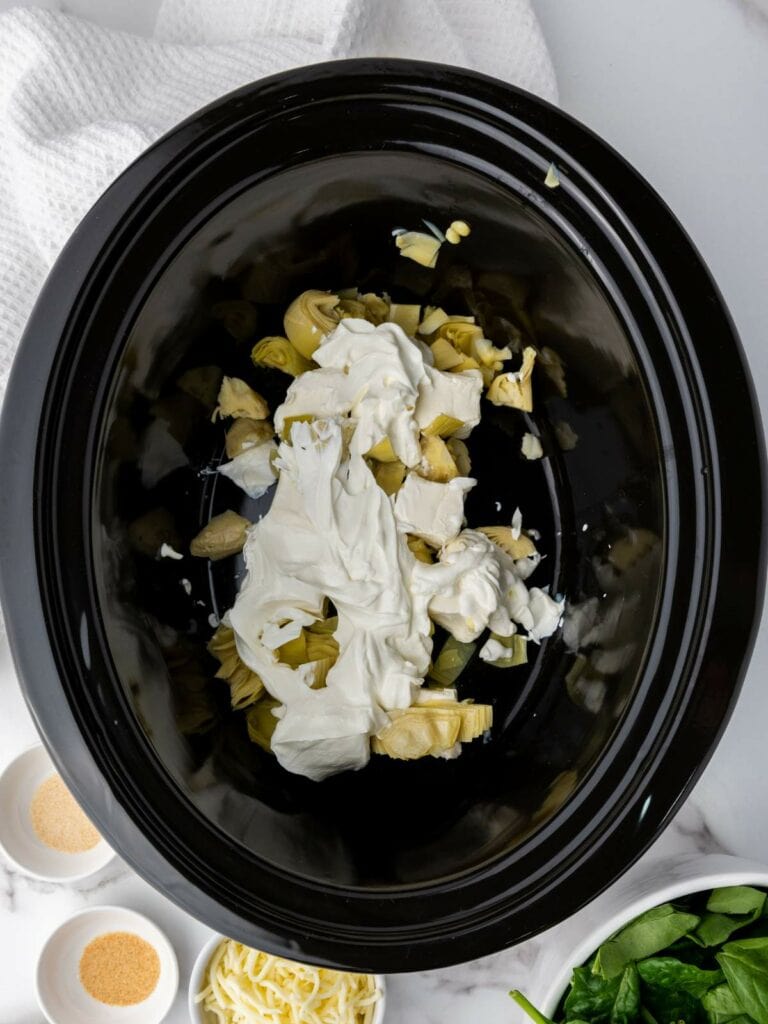 Ingredients including cream cheese and artichokes in a black slow cooker pot.