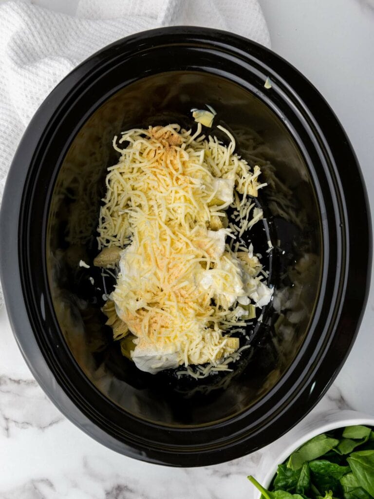 Grated cheese and cream cheese in a black slow cooker on a marble surface.