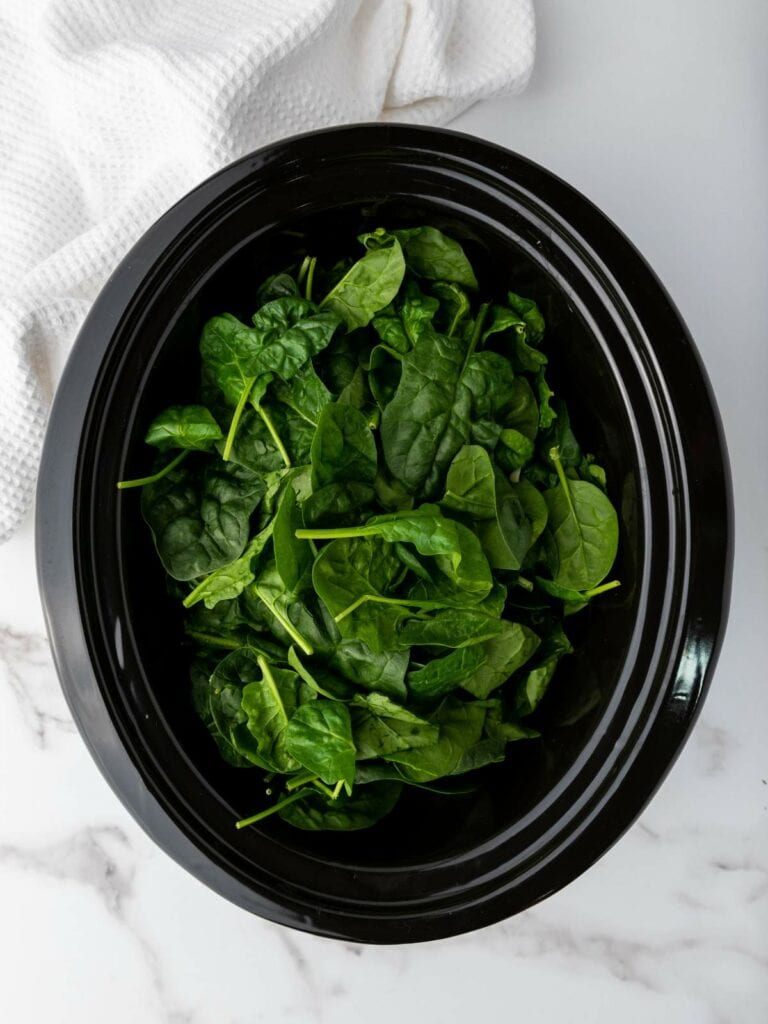 Fresh spinach leaves in a black bowl on a marble surface with a white cloth nearby.