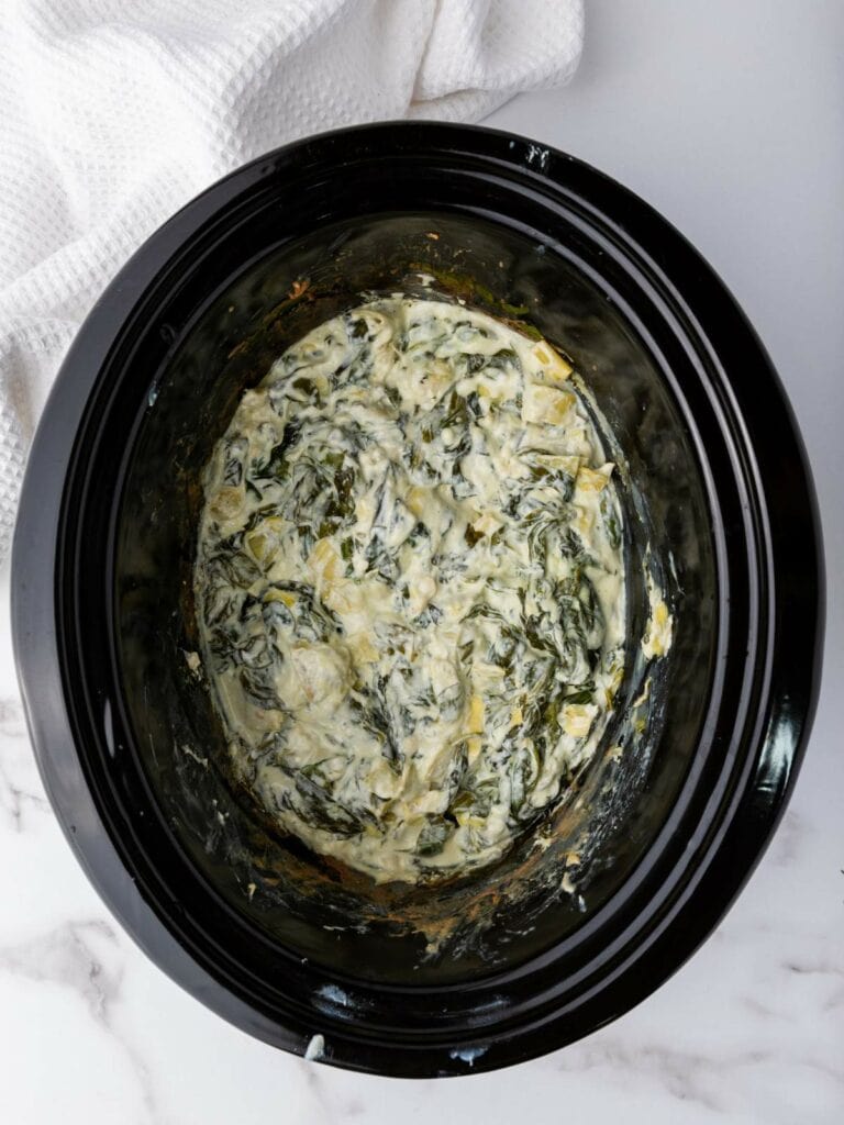 A slow cooker containing a creamy spinach and artichoke dip sits on a marble countertop next to a white towel.