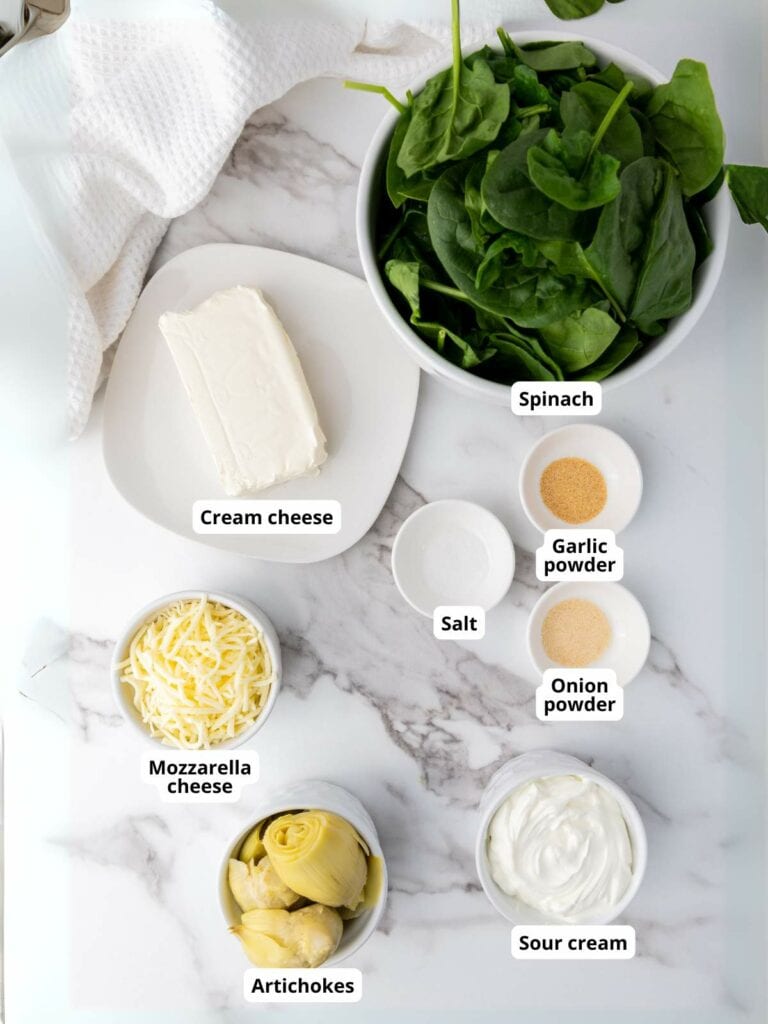 Ingredients for a spinach artichoke dip are arranged on a marble surface.