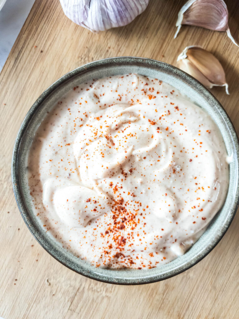 Bowl of spicy mayonnaise topped with red pepper flakes on a wooden surface, with garlic cloves nearby.