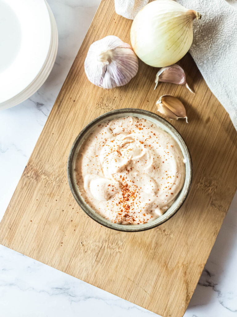 Bowl of mayonnaise sprinkled with red pepper, surrounded by a garlic bulb, cloves, an onion, and a towel.