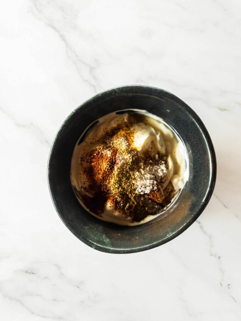 Black bowl with yogurt topped with various spices, placed on a white marbled surface.