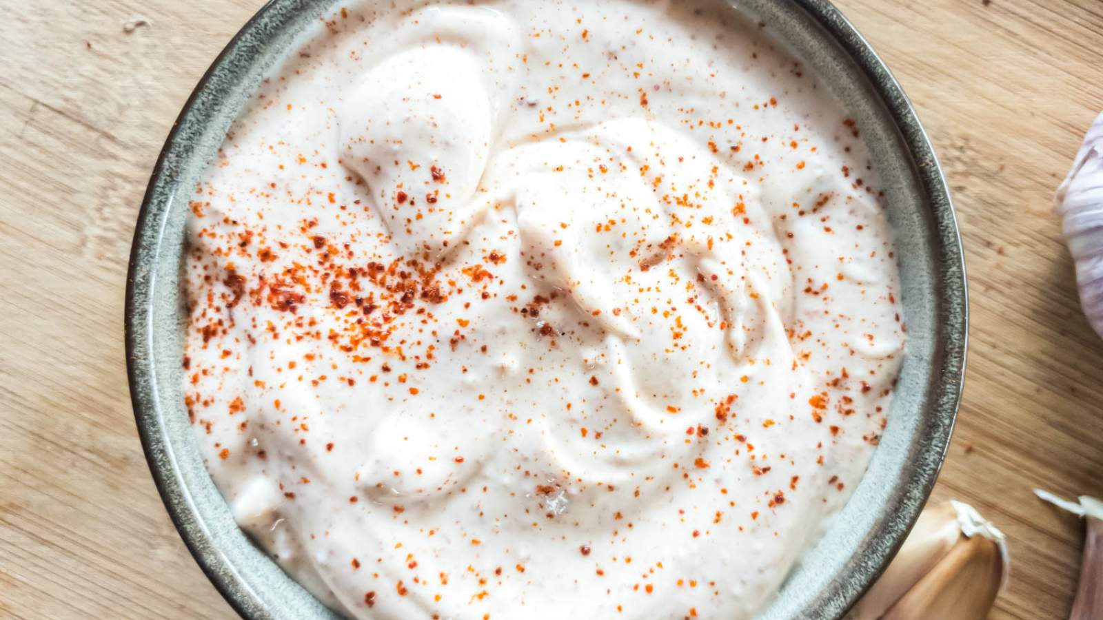 A bowl of spicy mayonnaise, sprinkled with red seasoning, on a wooden surface next to garlic cloves.