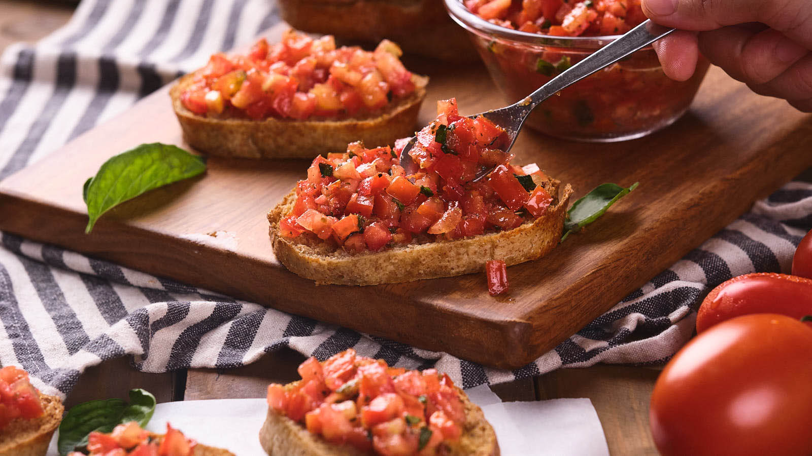 Tomato bruschetta on a wooden board with a hand adding more tomato mixture.