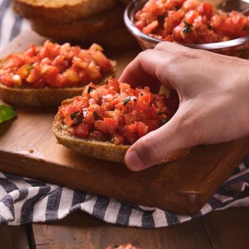 A hand is placing a slice of bread topped with diced tomatoes and herbs onto a wooden board.