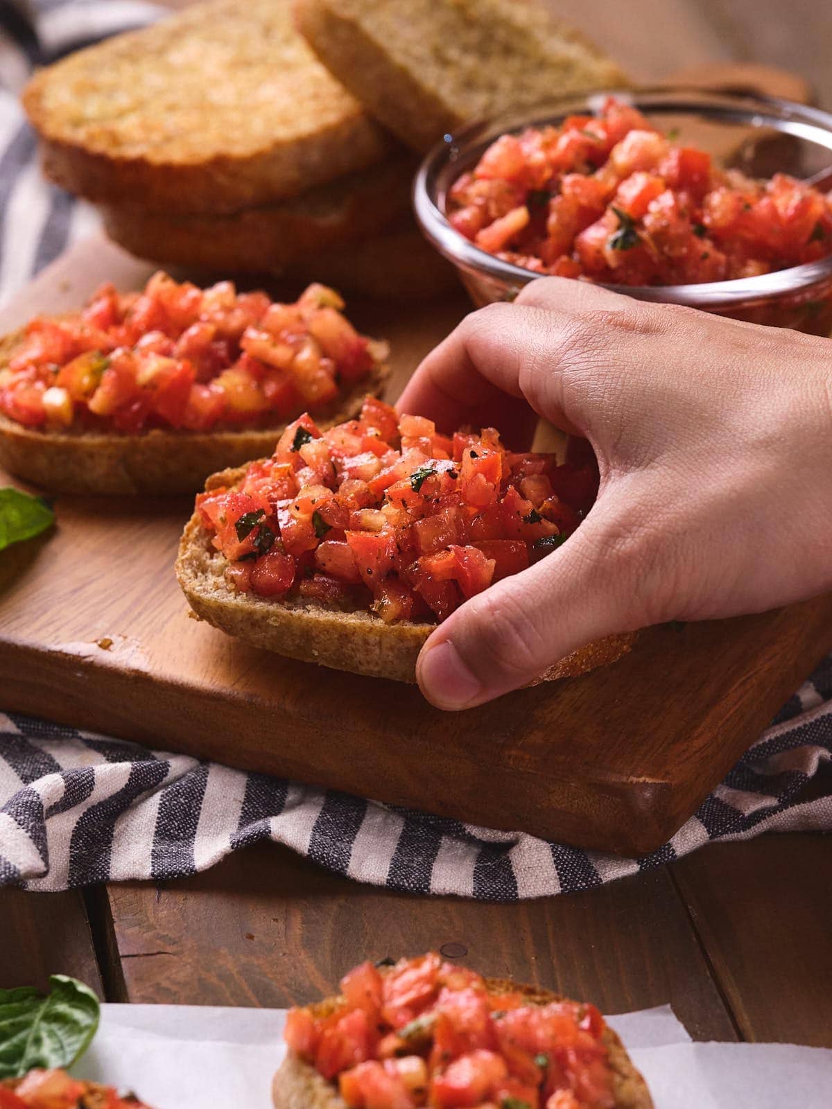 A hand is placing a slice of bread topped with diced tomatoes and herbs onto a wooden board.