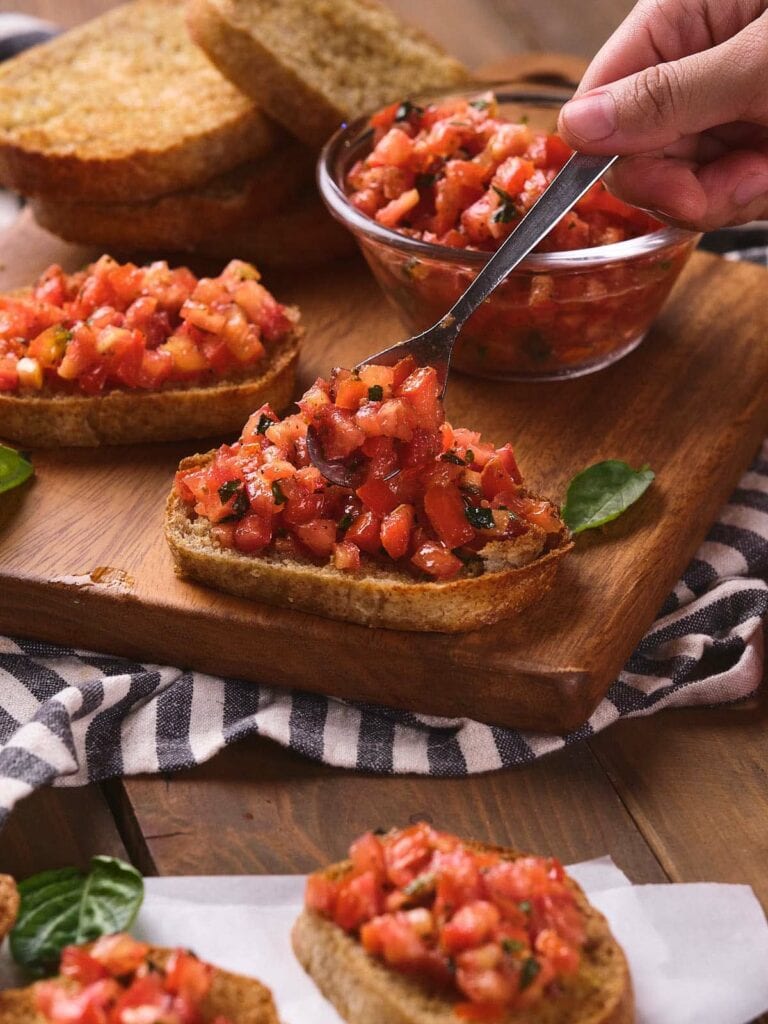 Hand placing tomato bruschetta topping on toasted bread.