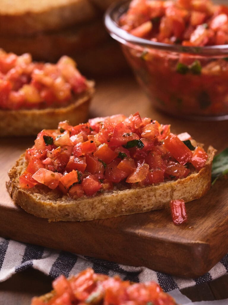 A slice of bread topped with diced tomatoes and herbs, with more servings and a bowl of the mixture in the background.