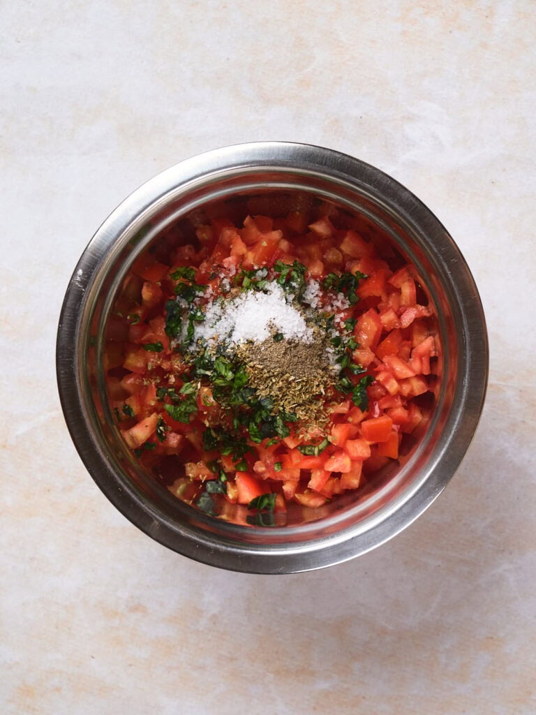 Chopped tomatoes, herbs, and spices in a stainless steel bowl on a light-colored surface.