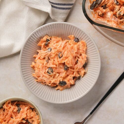 Three bowls of carrot salad with raisins and a creamy dressing, alongside a fork and a cloth napkin.