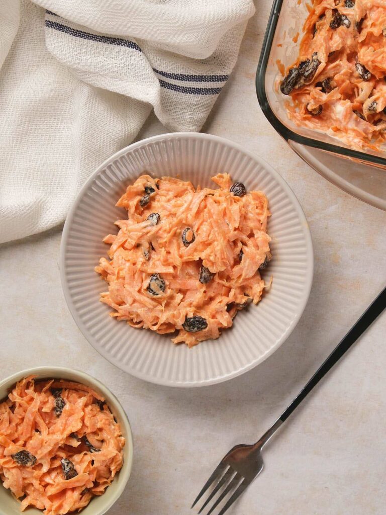 Three bowls of carrot salad with raisins and a creamy dressing, alongside a fork and a cloth napkin.