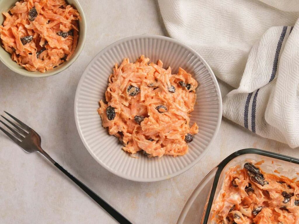 A bowl of carrot salad with raisins on a textured surface,.