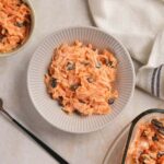 A bowl of carrot salad with raisins on a textured surface,.