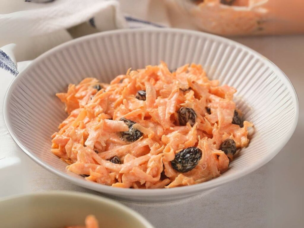 A bowl of carrot salad with raisins in a white ribbed dish.