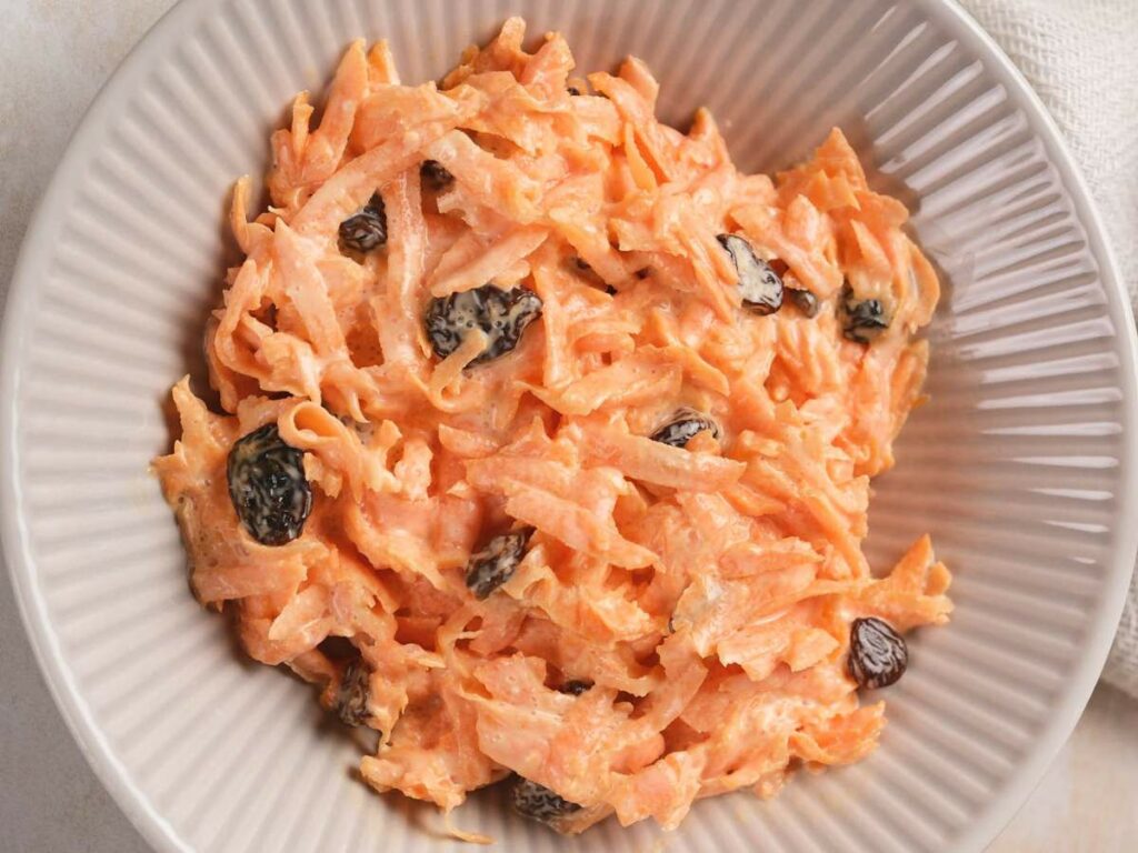 A bowl of carrot salad mixed with raisins in a ridged, round, white dish.