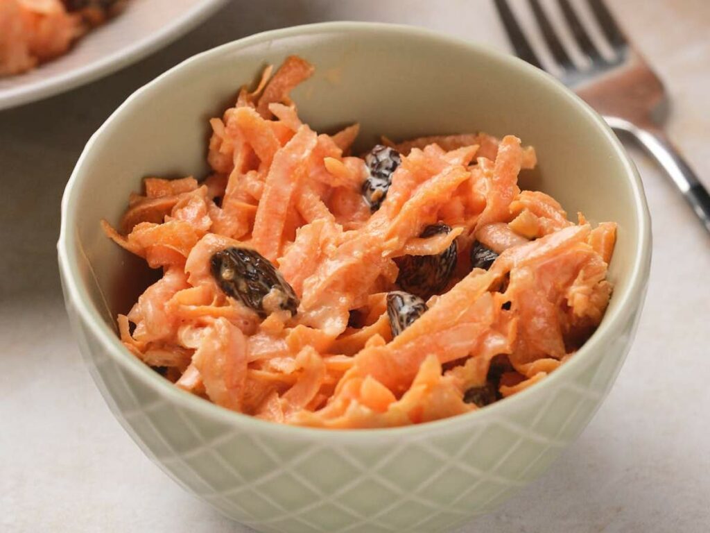 A bowl of carrot salad with raisins, served in a textured bowl, placed on a light-colored surface beside a fork.