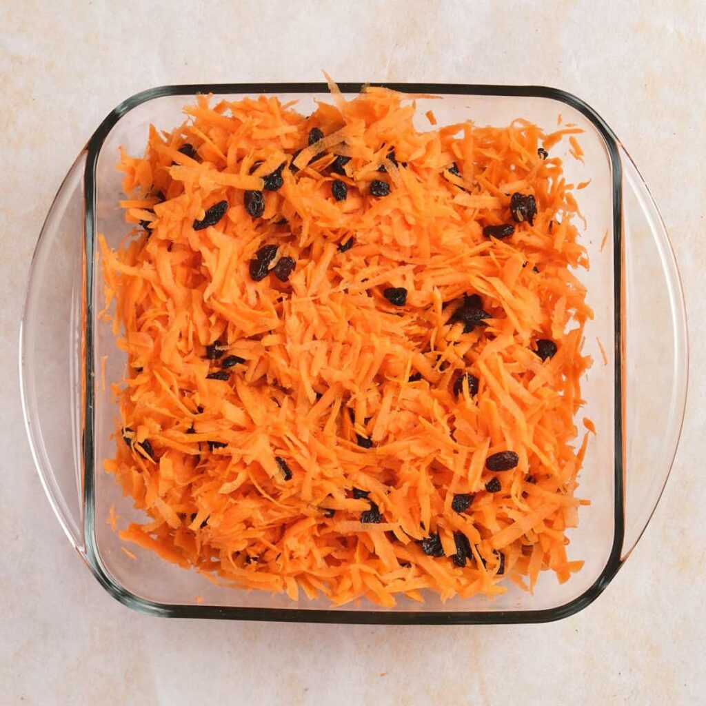 Grated carrots mixed with raisins in a square glass baking dish on a light surface.