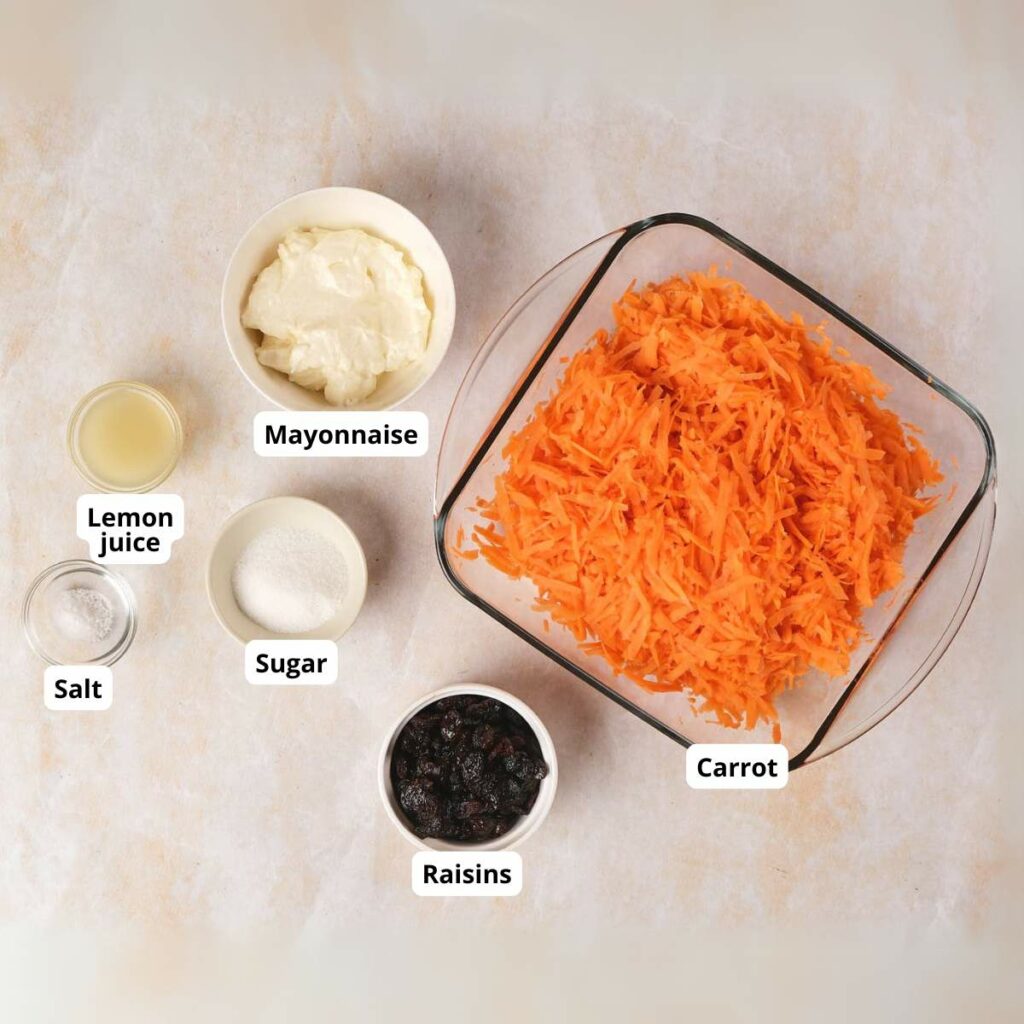 Ingredients for carrot salad: grated carrot in a bowl, small bowls of mayonnaise, raisins, lemon juice, sugar, and salt.